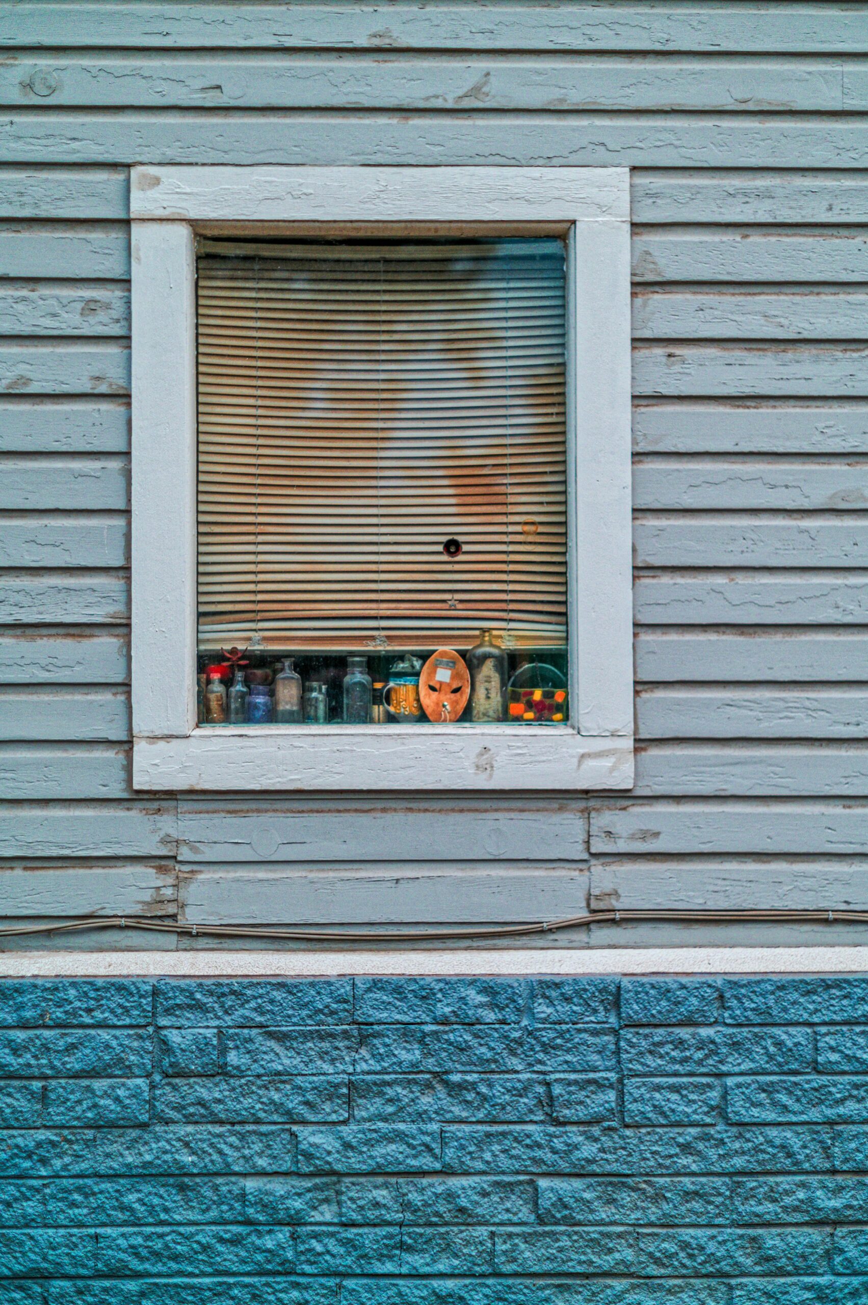 A vintage window with blinds partially open, showcasing jars and a clock.